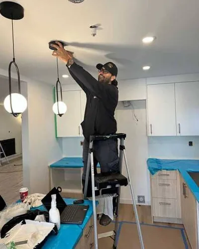 A Two Lions electrician on a step ladder setting a pendant light in a kitchen that is still under renovation, cabinets in and counters still protected.